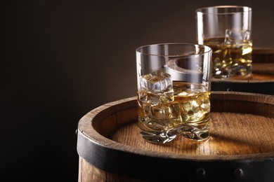 Photo of Whiskey with ice cubes in glasses on wooden barrels against dark background, closeup. Space for text