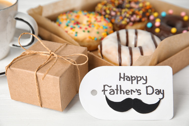 Photo of Tag with phrase HAPPY FATHER'S DAY, donuts, coffee and gift on white wooden table