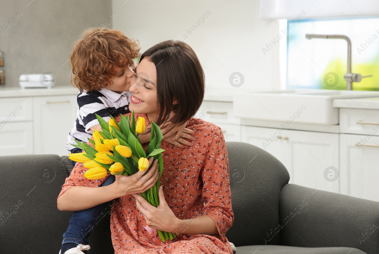 Photo of Little son congratulating his mom with Mother`s day at home, space for text. Woman holding bouquet of yellow tulips