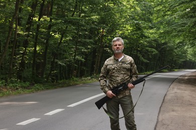 Photo of Man wearing camouflage with hunting rifle near road in forest. Space for text