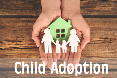 Image of Woman holding figures of family and green house in hands on wooden background, top view. Child adoption concept