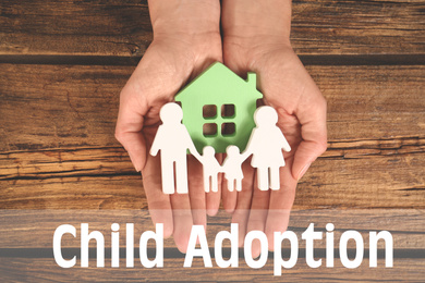 Woman holding figures of family and green house in hands on wooden background, top view. Child adoption concept