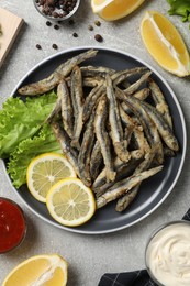 Delicious fried anchovies with lemon, lettuce leaves and sauces served on light grey table, flat lay