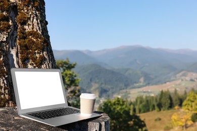 Modern laptop with blank screen and coffee cup on tree stump in mountains, space for text. Working outdoors