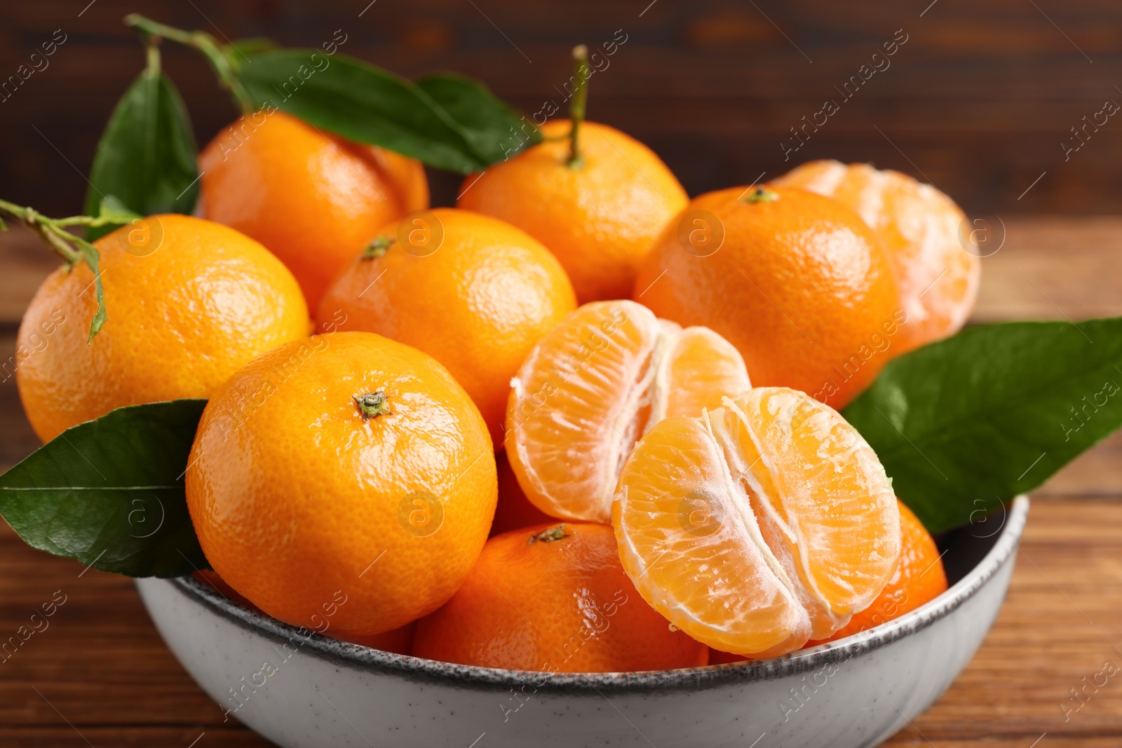 Photo of Fresh tangerines with green leaves in bowl on wooden table, closeup