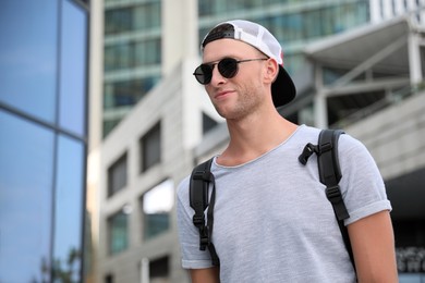 Photo of Handsome young man with stylish sunglasses and backpack near building outdoors, space for text
