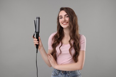 Photo of Happy young woman with beautiful hair holding curling iron on grey background