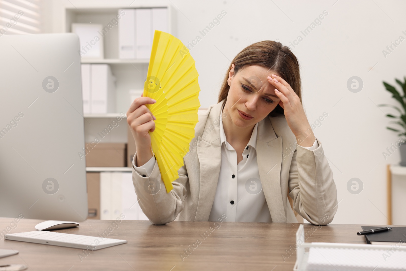 Photo of Businesswoman waving yellow hand fan to cool herself at table in office