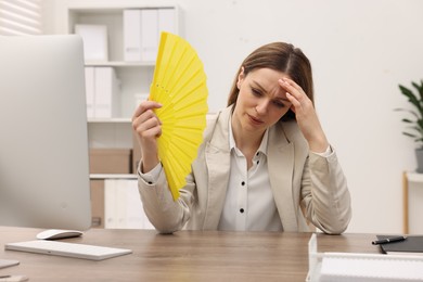 Businesswoman waving yellow hand fan to cool herself at table in office