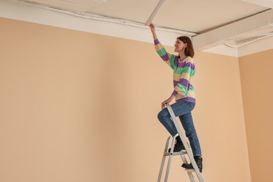 Photo of Young woman installing ceiling lamp on stepladder indoors, space for text