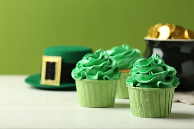 Photo of St. Patrick's day party. Tasty cupcakes with green cream, pot of gold and leprechaun hat on white table, closeup. Space for text