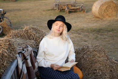 Photo of Beautiful woman with book sitting on wooden bench near hay bales outdoors. Autumn season