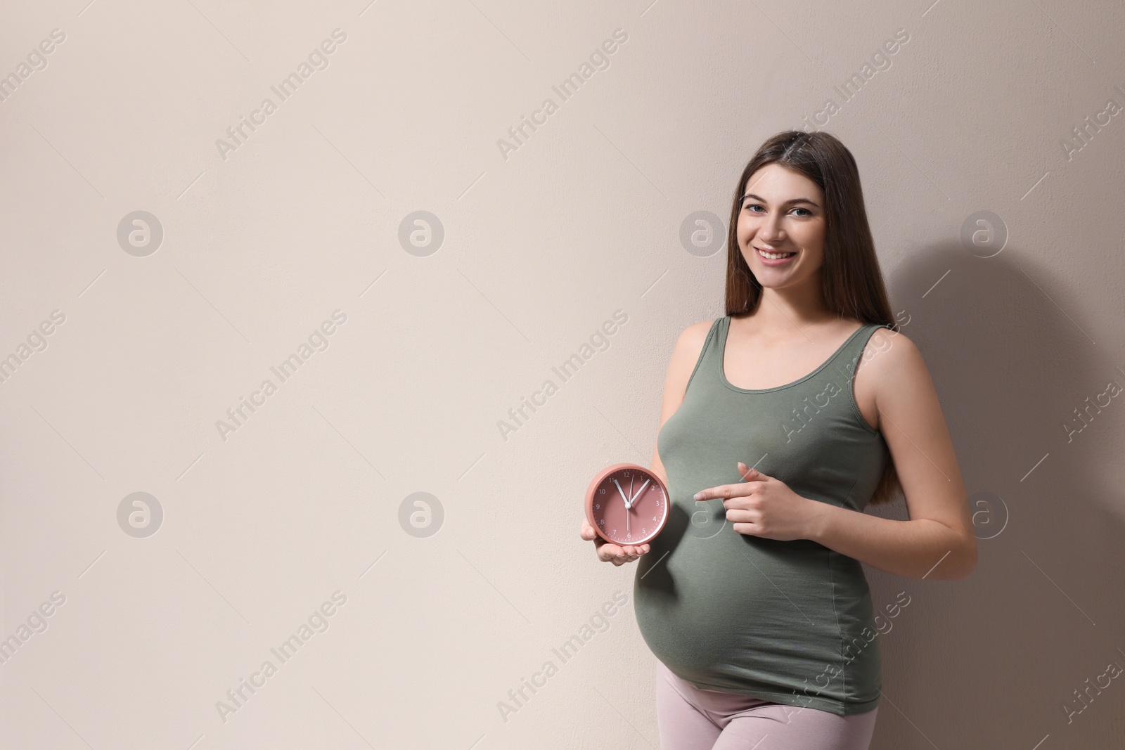 Photo of Young pregnant woman pointing at clock near her belly on beige background, space for text. Time to give birth