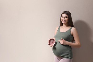 Young pregnant woman pointing at clock near her belly on beige background, space for text. Time to give birth