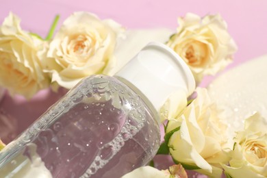 Photo of Wet bottle of micellar water and beautiful white roses on pink background, closeup