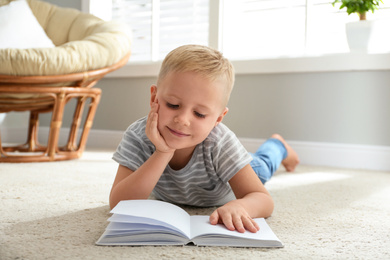 Photo of Cute little boy reading book on floor at home