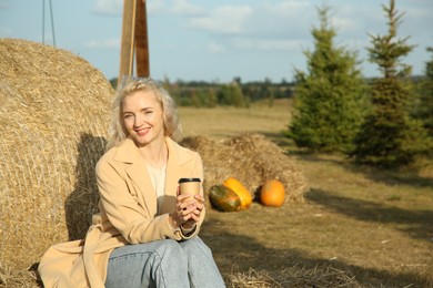 Photo of Beautiful woman with cup of hot drink sitting near hay bale outdoors, space for text. Autumn season