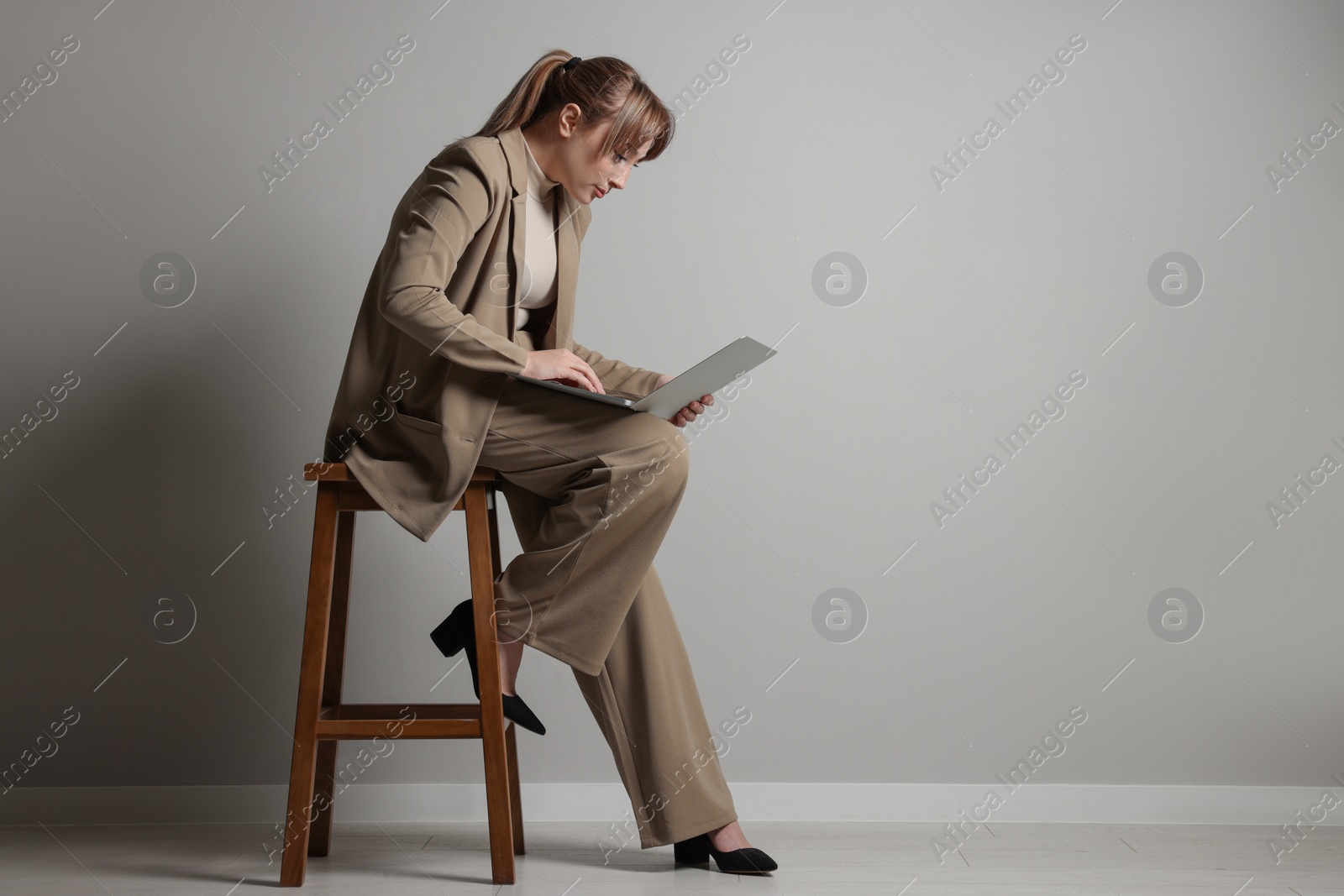 Photo of Young woman with poor posture using laptop while sitting on stool near grey wall, space for text