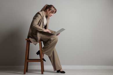 Photo of Young woman with poor posture using laptop while sitting on stool near grey wall, space for text