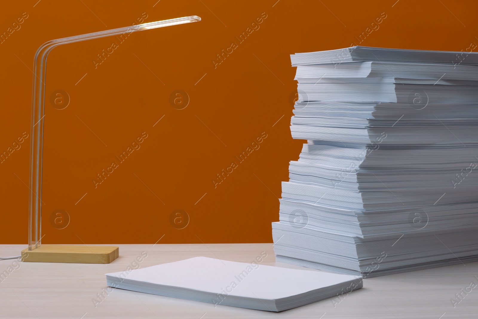 Photo of Stack of paper sheets and lamp on white wooden table near orange wall