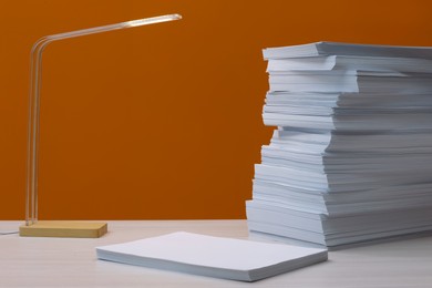 Stack of paper sheets and lamp on white wooden table near orange wall