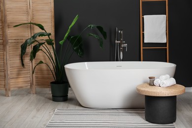 Photo of Spa day. Ceramic tub, houseplant and wicker table in bathroom