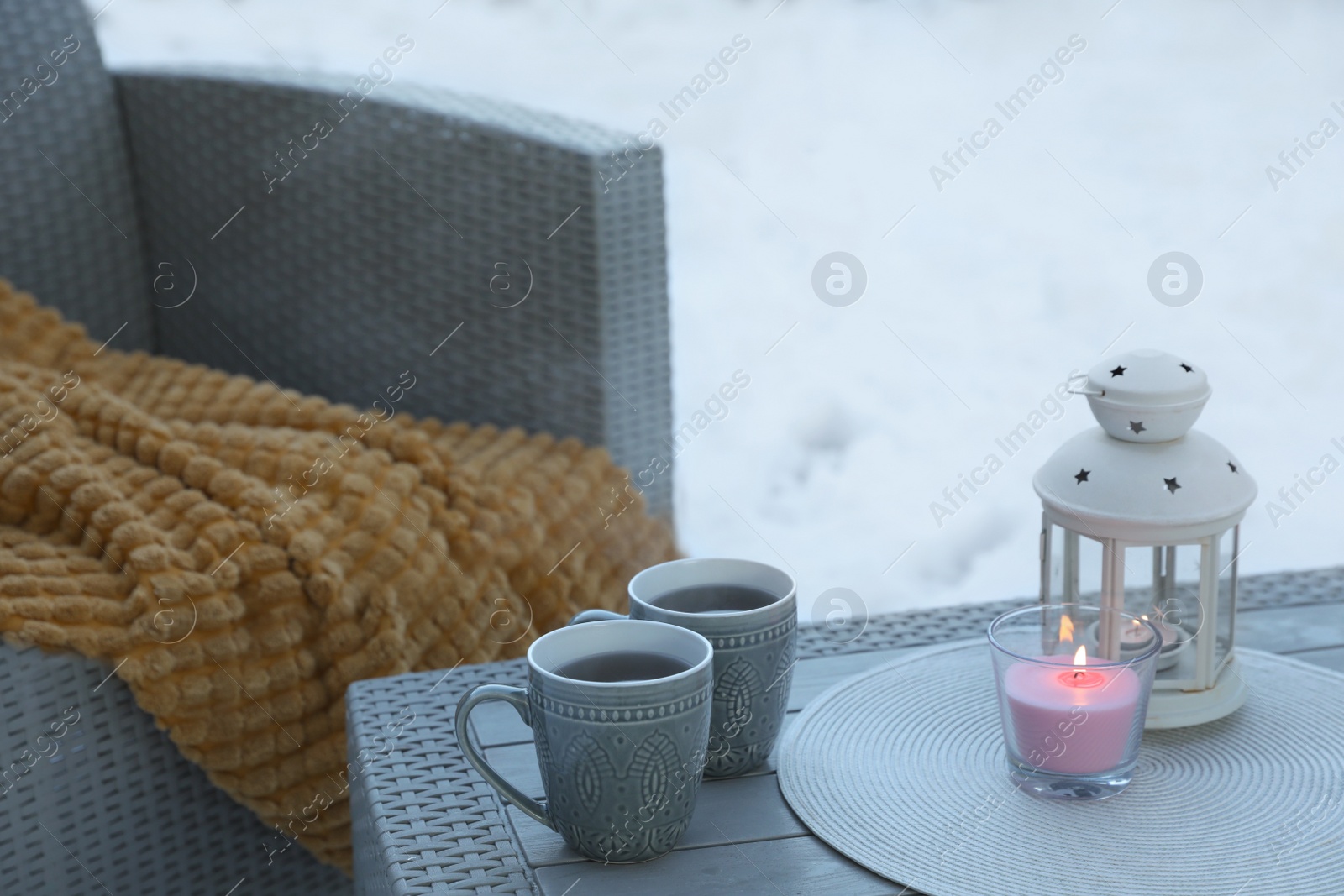 Photo of Burning candle, lantern and cups with hot drink on coffee table outdoors. Cosy winter