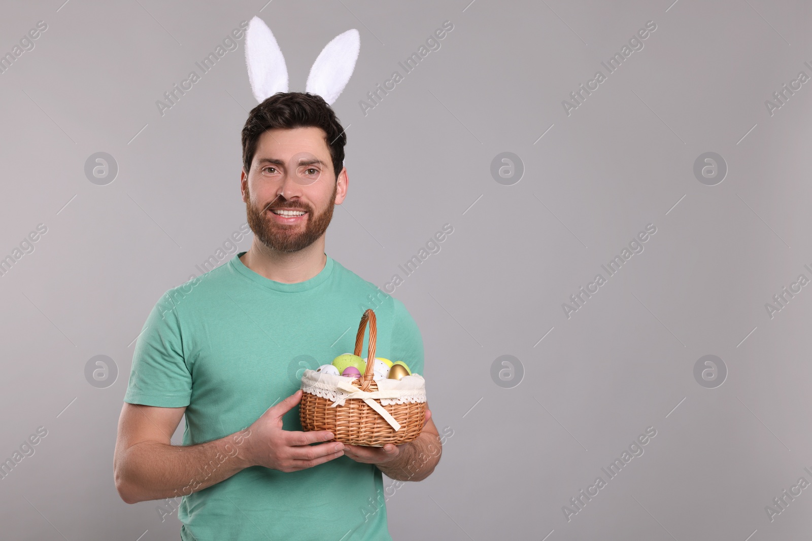 Photo of Portrait of happy man in cute bunny ears headband holding wicker basket with Easter eggs on light grey background. Space for text