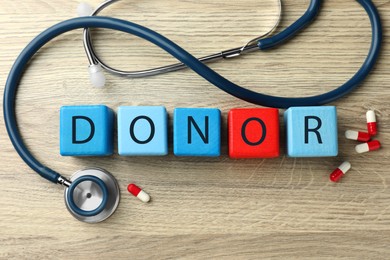 Word Donor made of cubes, stethoscope and pills on wooden table, flat lay