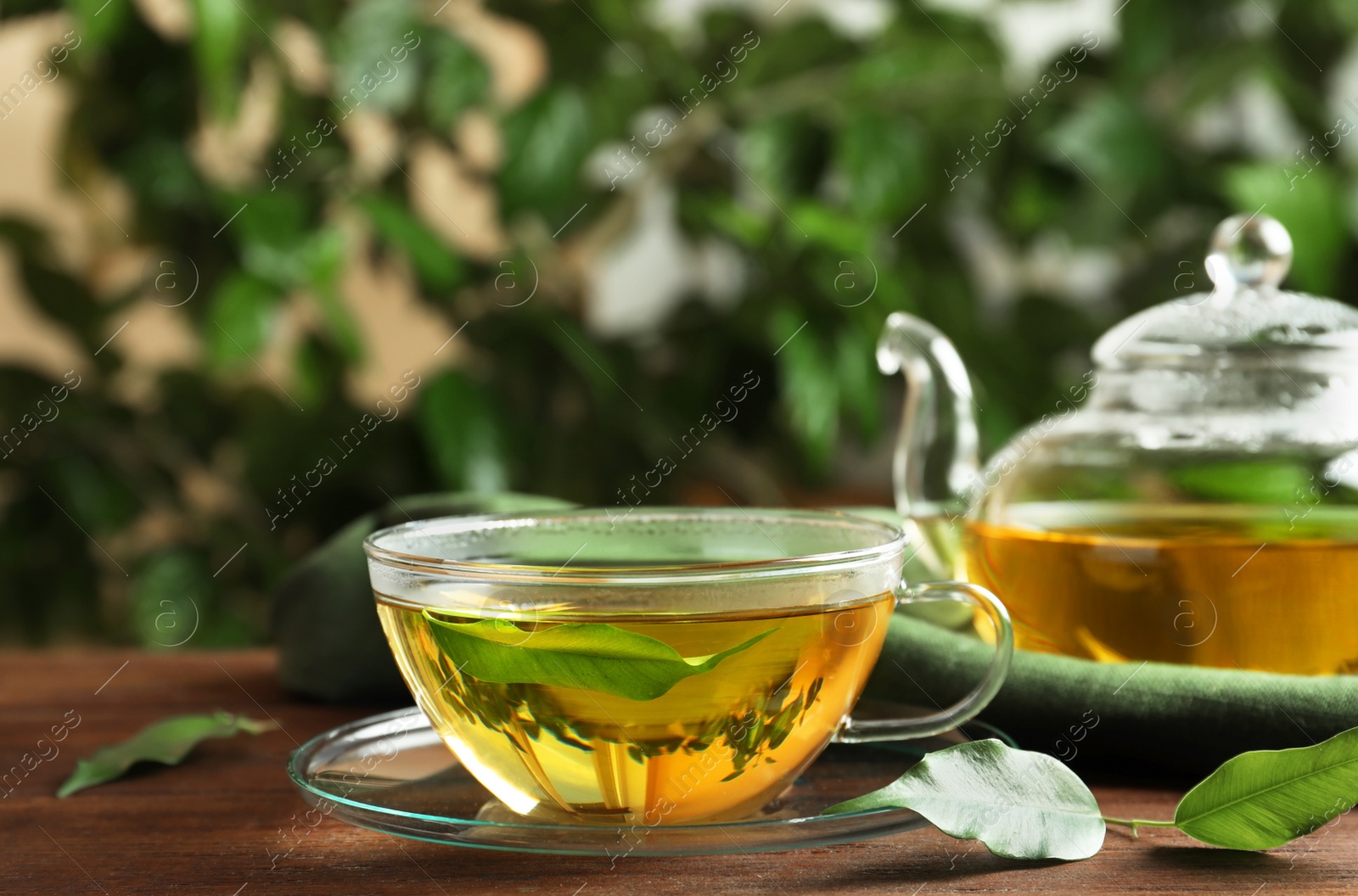 Photo of Fresh green tea in glass cup with saucer, leaves and teapot on wooden table