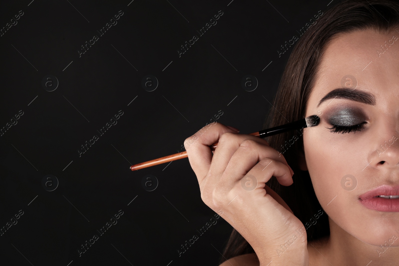Photo of Portrait of beautiful woman applying stylish makeup with brush against dark background. Space for text