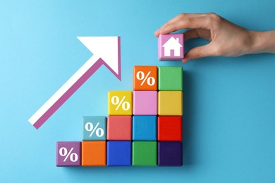Mortgage rate rising illustrated by upward arrow. Woman putting cube with house icon near other ones on light blue background, top view