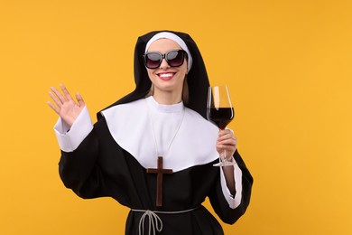 Photo of Happy woman in nun habit and sunglasses holding glass of wine against orange background. Sexy costume