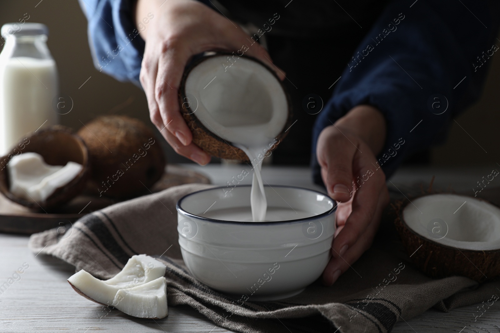 Photo of Woman pouring coconut milk into bowl at white wooden table, closeup