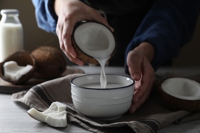 Woman pouring coconut milk into bowl at white wooden table, closeup