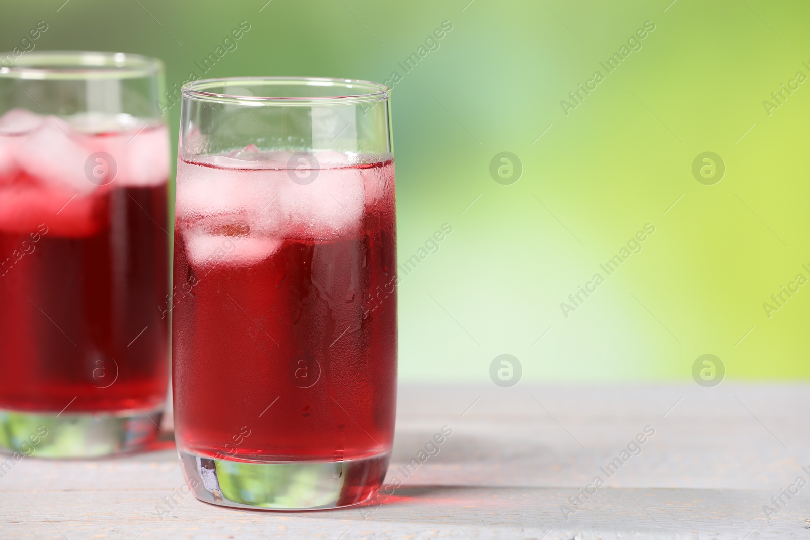 Photo of Refreshing hibiscus tea with ice cubes in glasses on white wooden table against blurred green background, closeup. Space for text