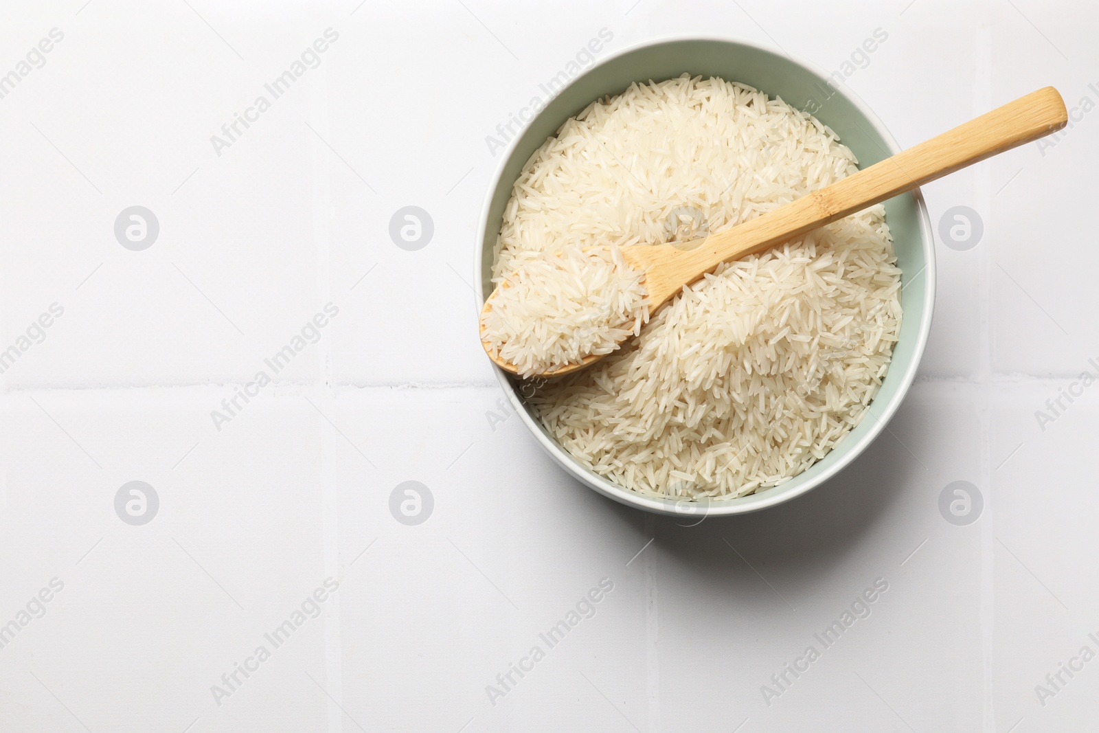 Photo of Raw basmati rice in bowl and spoon on white tiled table, top view. Space for text