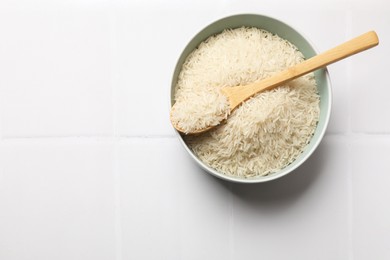 Photo of Raw basmati rice in bowl and spoon on white tiled table, top view. Space for text