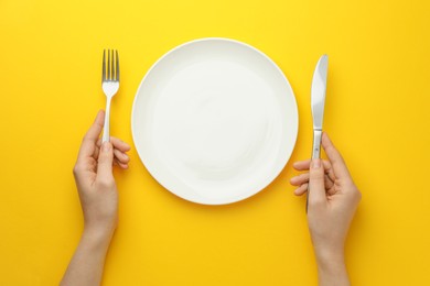 Photo of Woman with fork, knife and clean plate at yellow table, top view