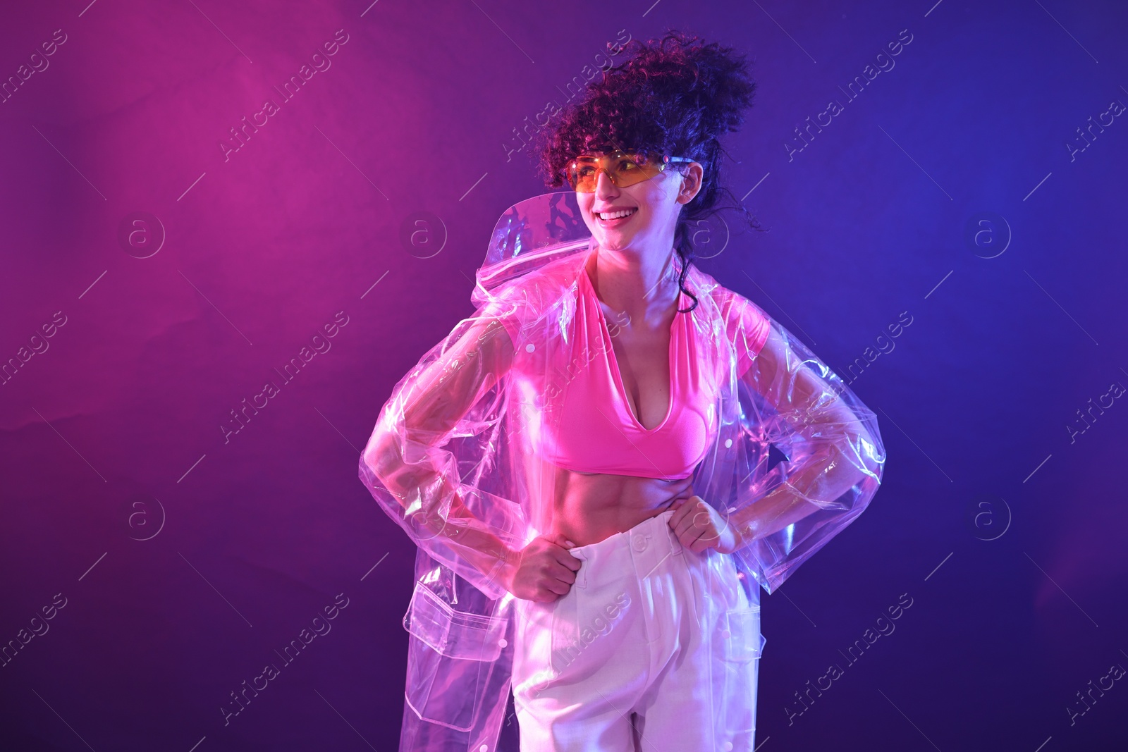 Photo of Beautiful young woman in transparent coat and sunglasses posing on color background in neon lights