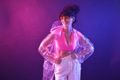 Photo of Beautiful young woman in transparent coat and sunglasses posing on color background in neon lights