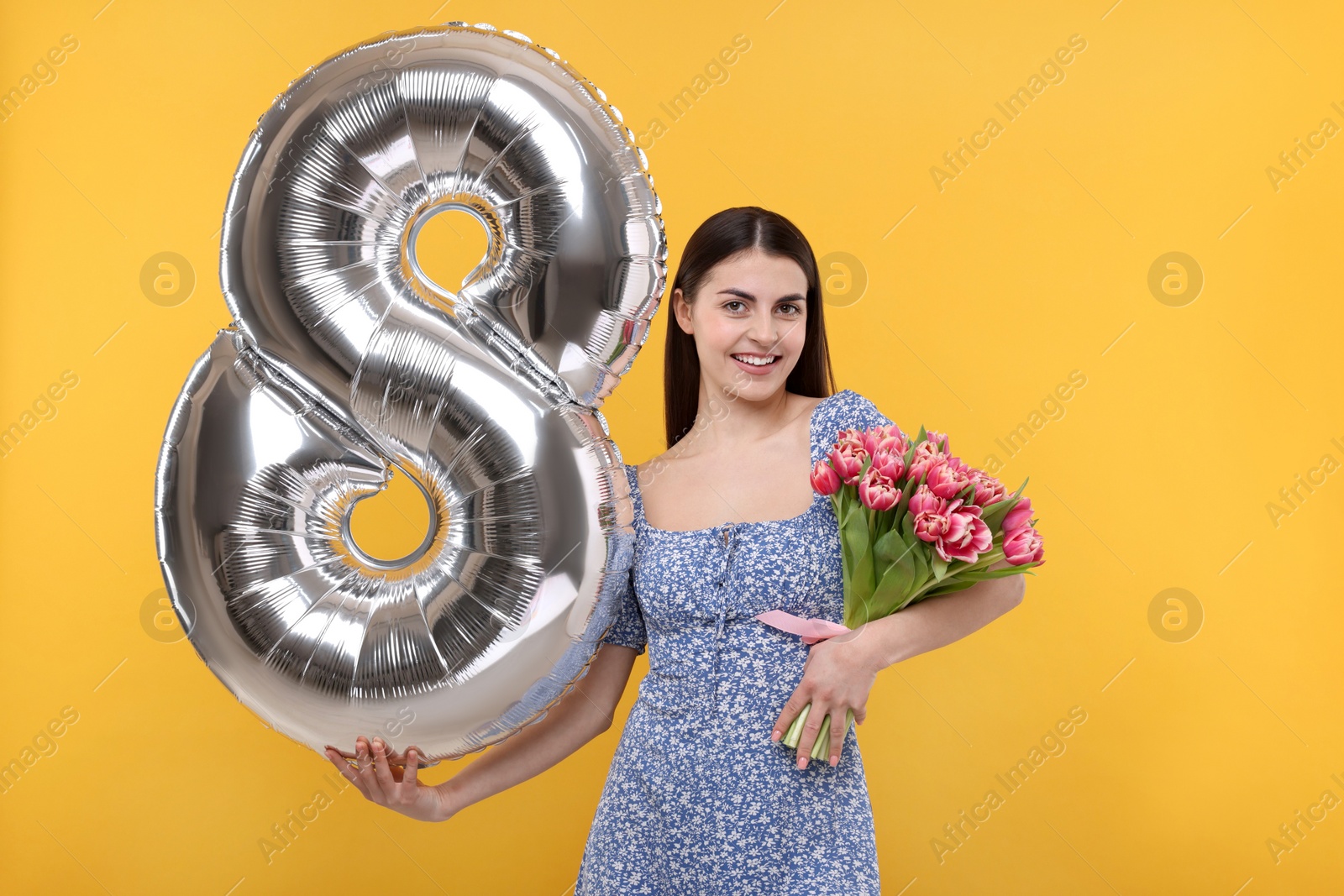 Photo of Happy Women's Day. Charming lady holding bouquet of beautiful flowers and balloon in shape of number 8 on orange background