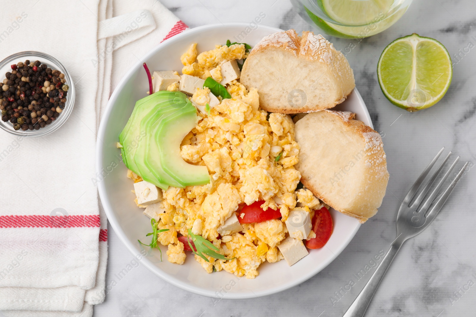 Photo of Bowl with delicious scrambled eggs, tofu, avocado and slices of baguette on white marble table, flat lay