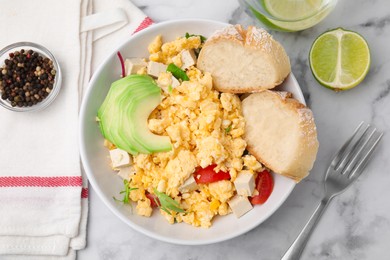 Bowl with delicious scrambled eggs, tofu, avocado and slices of baguette on white marble table, flat lay