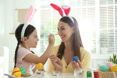 Photo of Happy mother and daughter with bunny ears headbands having fun while painting Easter eggs at home