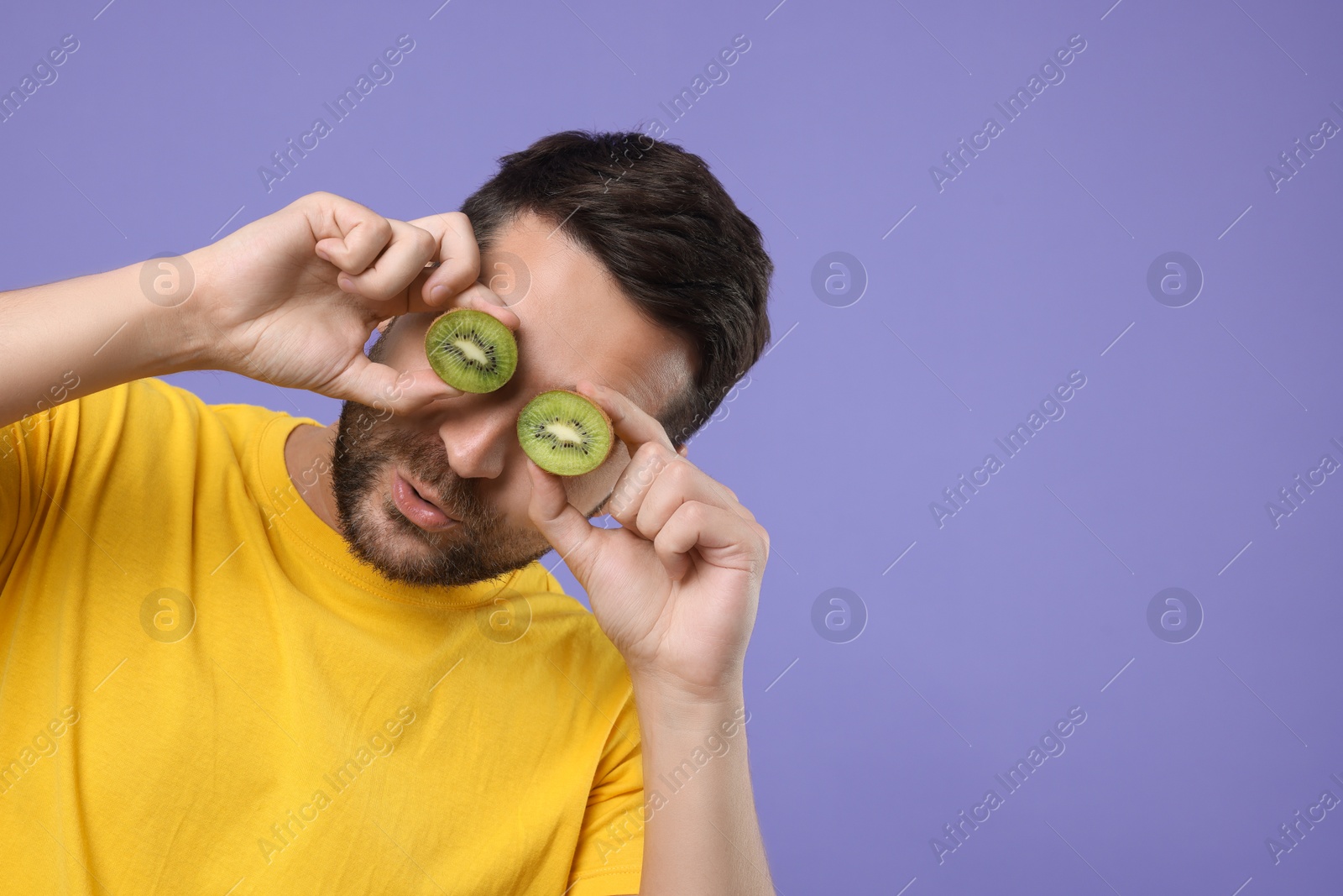 Photo of Man covering his eyes with halves of kiwi on purple background. Space for text