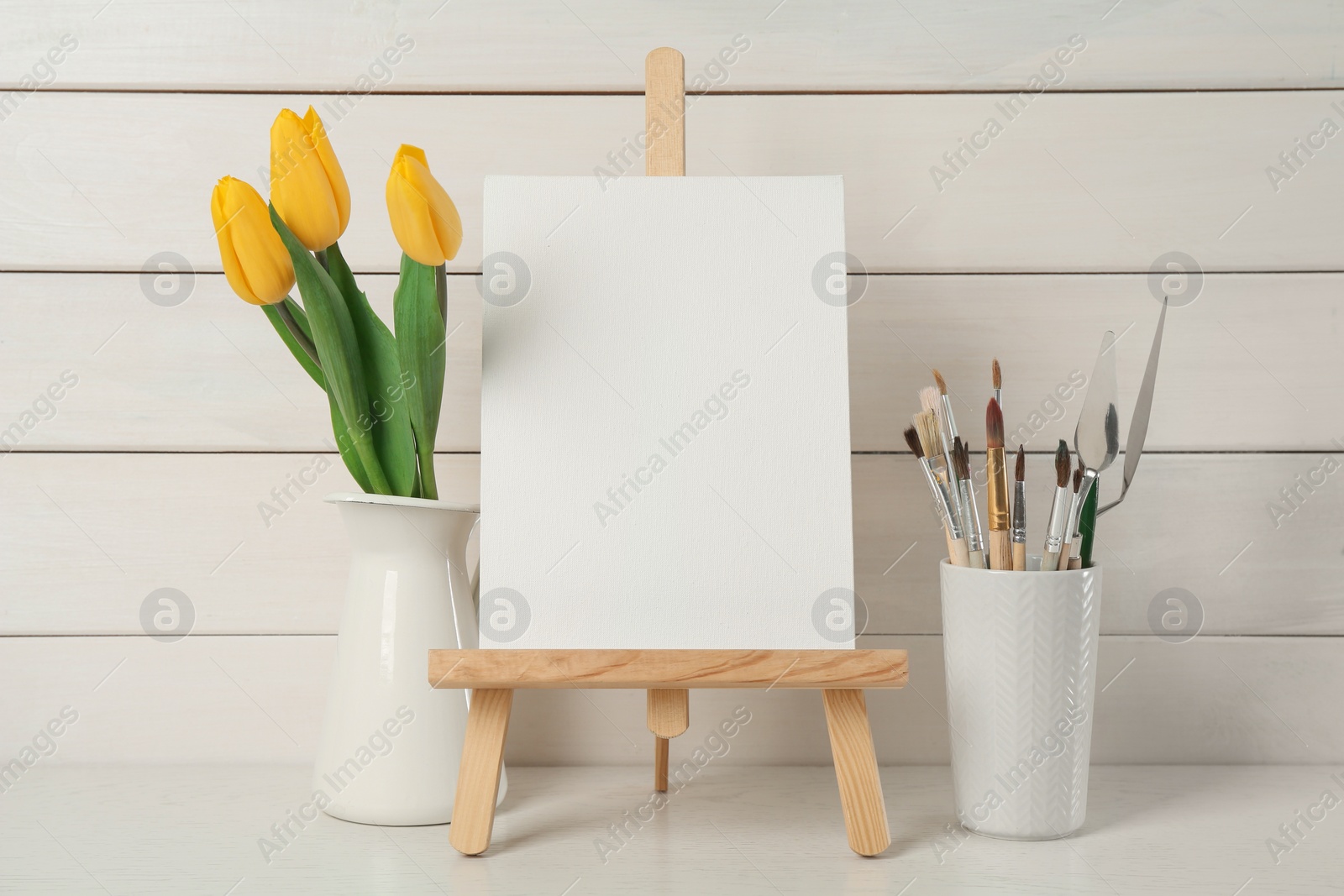 Photo of Easel with blank canvas, brushes and tulips on white wooden table