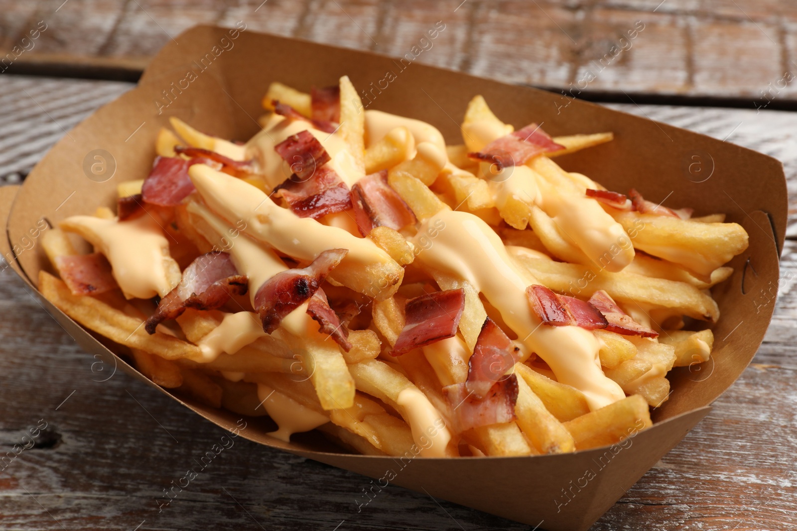 Photo of Tasty potato fries, cheese sauce and bacon in paper container on wooden table, closeup