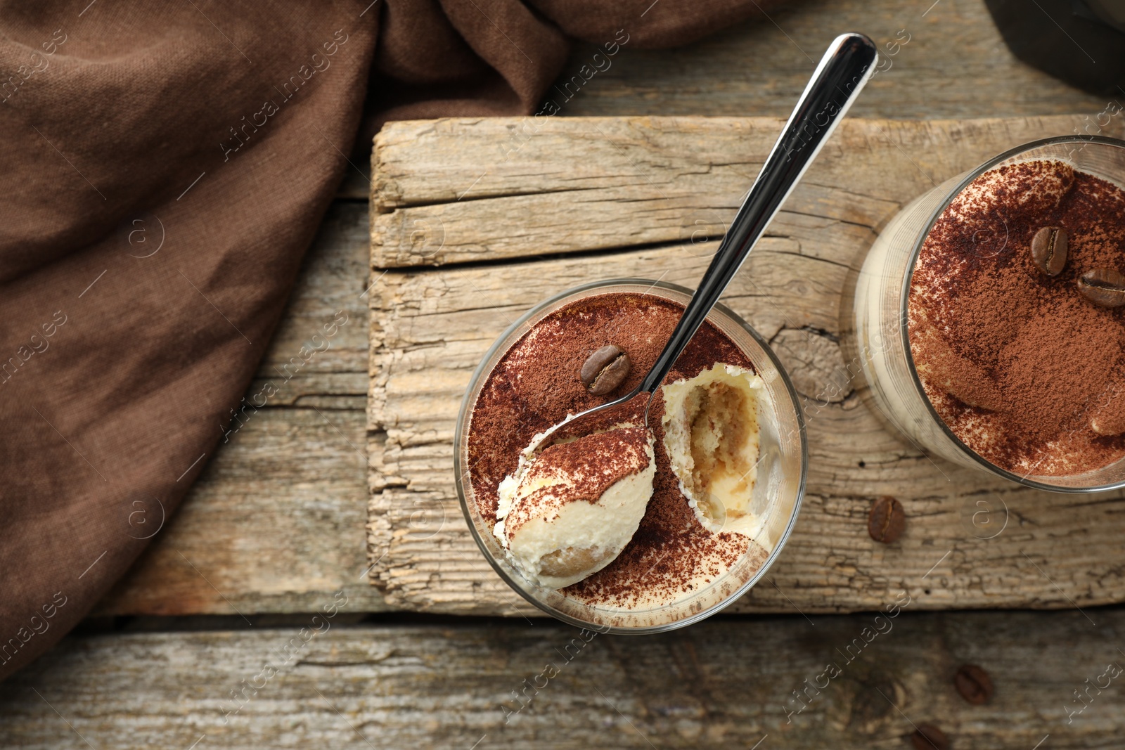 Photo of Delicious tiramisu in glasses, spoon and coffee beans on wooden table, top view