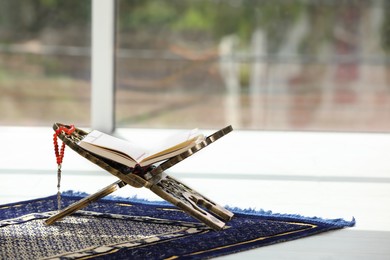 Rehal with open Quran and Misbaha on Muslim prayer rug near window indoors, space for text
